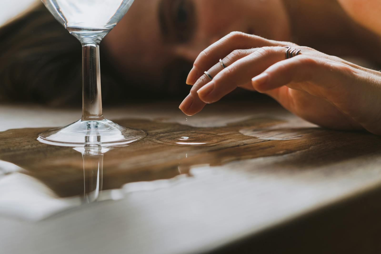 A close-up shot of a hand touching a water droplet next to a glass on a wooden surface.